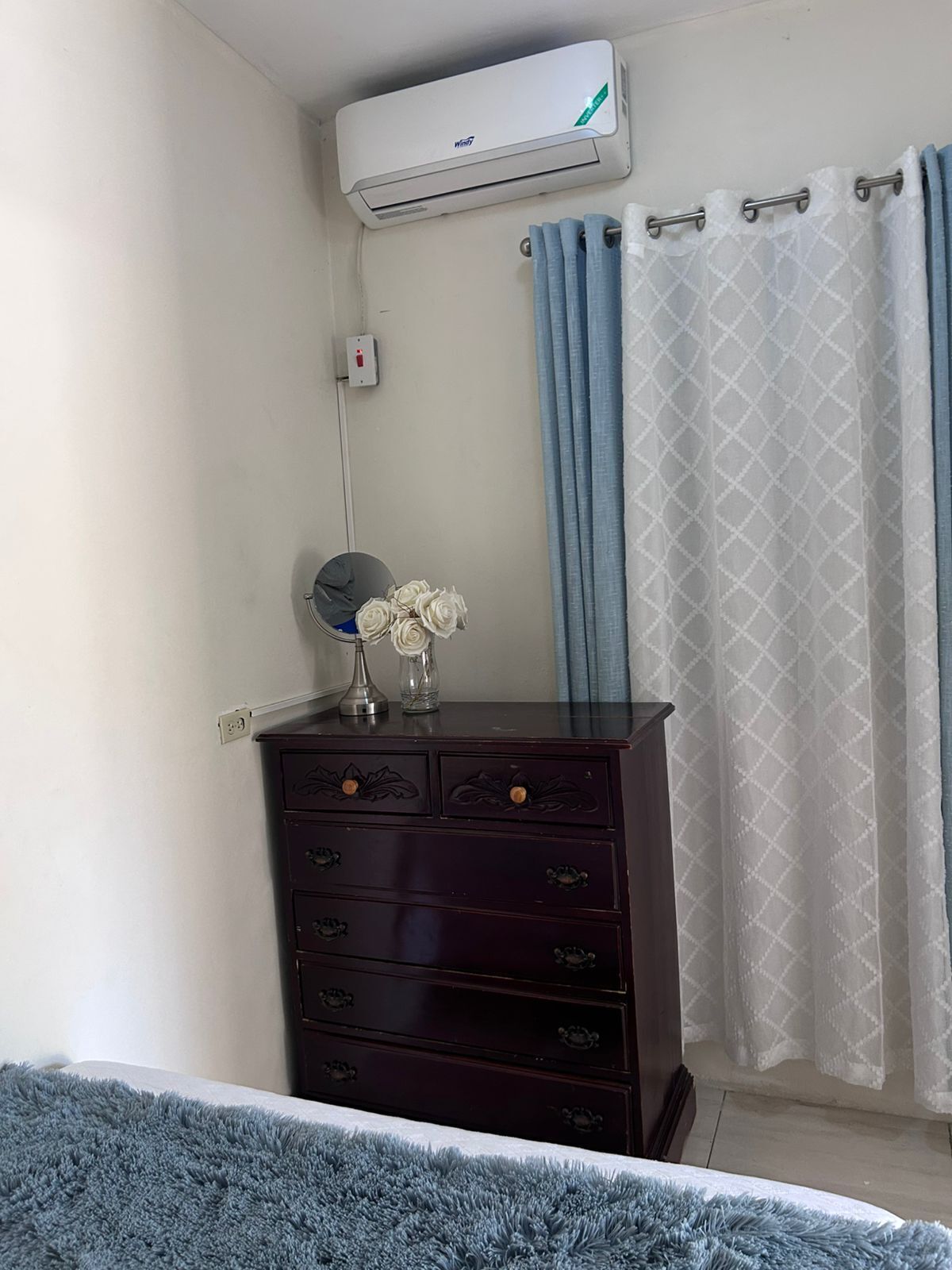 Een slaapkamer met een donker houten dressoir, een wandairconditioner, een stopcontact, een vaas met witte bloemen en een badgordijn met een blauw-wit patroon.