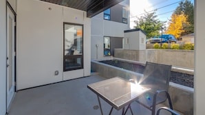 A modern outdoor patio with a glass table and chairs, a concrete planter, and a view of a residential street. - Fireplace, Courtyard, Free Heated Parking, Pets Ok (Calgary)