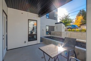 En modern uteplats med ett glasbord och stolar, en planteringslåda i betong och utsikt över en bostadsgata.