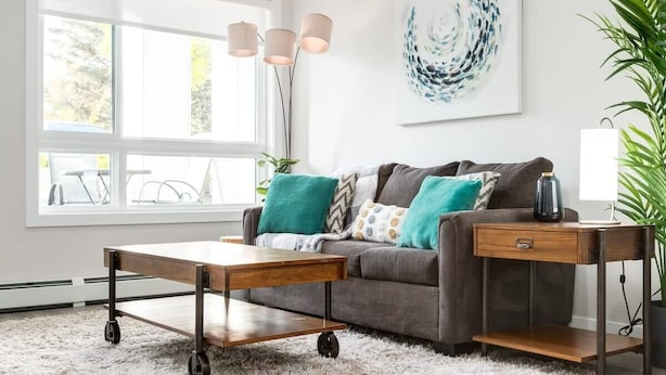 A modern living room with a sofa, a coffee table with wheels, a side table, and a large window with natural light. - Fireplace, Courtyard, Free Heated Parking, Pets Ok (Calgary)