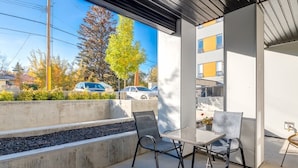 A modern outdoor patio with two metal chairs and a table, overlooking a parking area and buildings. - Fireplace, Courtyard, Free Heated Parking, Pets Ok (Calgary)