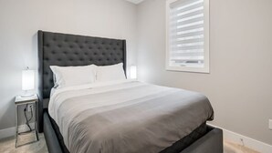 Ett sovrum med en stor säng, två sänglampor, ett fönster med persienner och ett vägguttag.