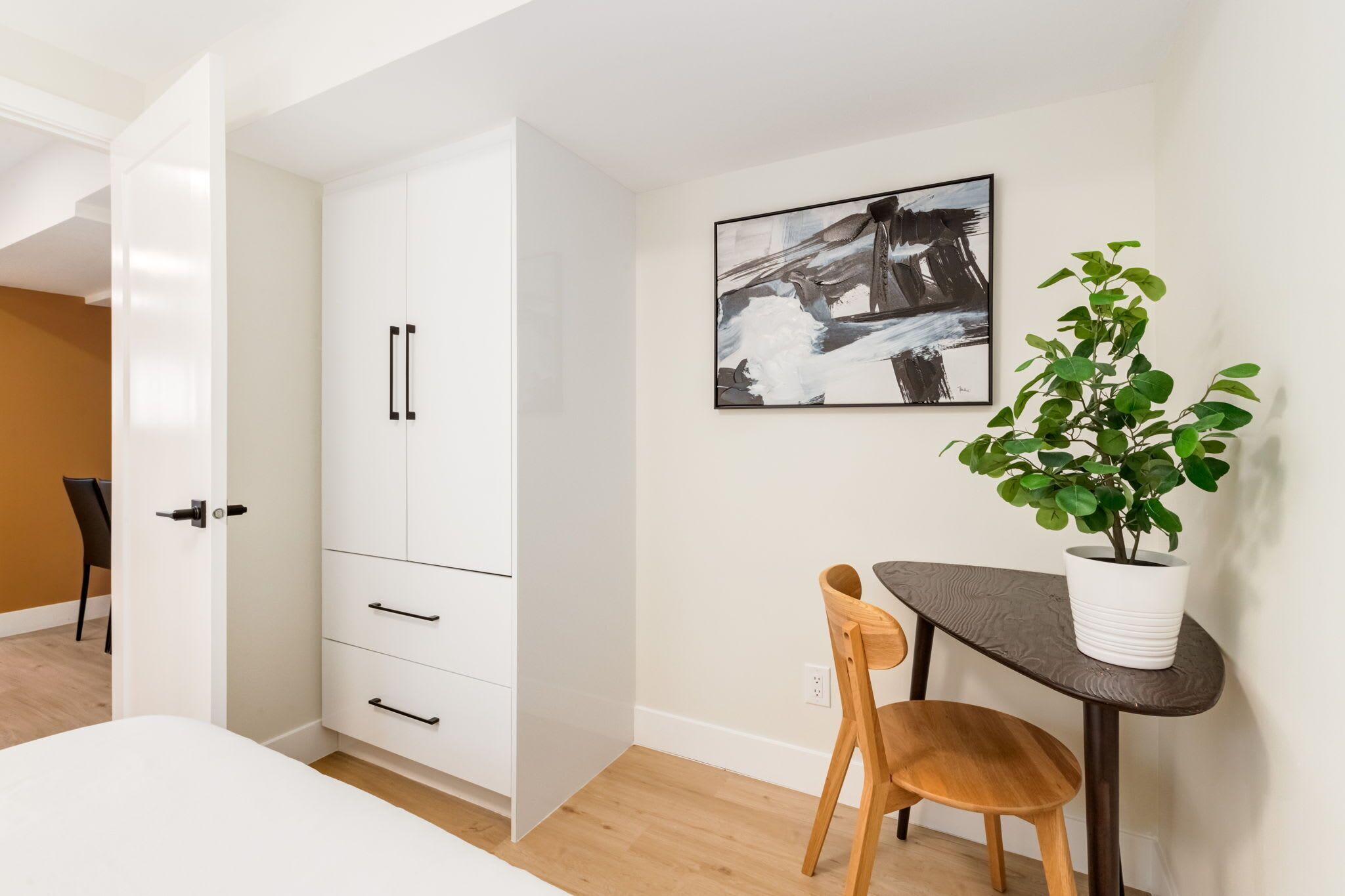 A modern room with a white wardrobe, a dining table with chairs, a potted plant, and a framed abstract painting on the wall.