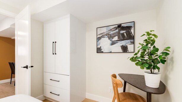 A modern room with a white wardrobe, a dining table with chairs, a potted plant, and a framed abstract painting on the wall.