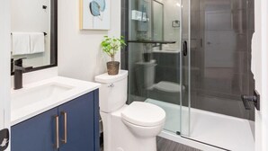 A modern bathroom with a glass shower enclosure, a white toilet, and a blue vanity with drawers. - Rare Find in Best Location, Work/Family Friendly (Calgary)