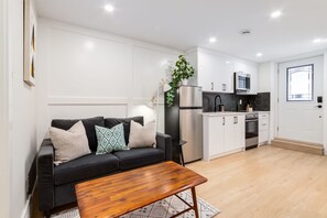 A modern living room with a black sofa, wooden coffee table, and a kitchen area with white cabinets and stainless steel appliances.