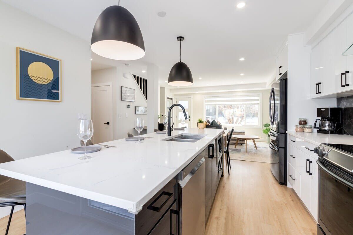 A modern kitchen with a central island, black pendant lights, and stainless steel appliances.