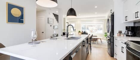 A modern kitchen with a central island, black pendant lights, and stainless steel appliances.
