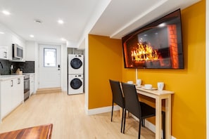 Una cocina moderna con armarios blancos, microondas y lavadora. Hay una zona de comedor con mesa y sillas, y un televisor grande montado en la pared que muestra una chimenea.