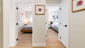 A hallway with two bedrooms, each with a bed, bedside tables, and framed artwork on the walls. - Rare Find in Best Location, Work/Family Friendly (Calgary)