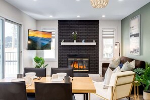 A modern living room with a fireplace, a dining area with a table and chairs, and a sofa. - Pet Friendly 4BDR w/ Main Floor office/Bed (Calgary)