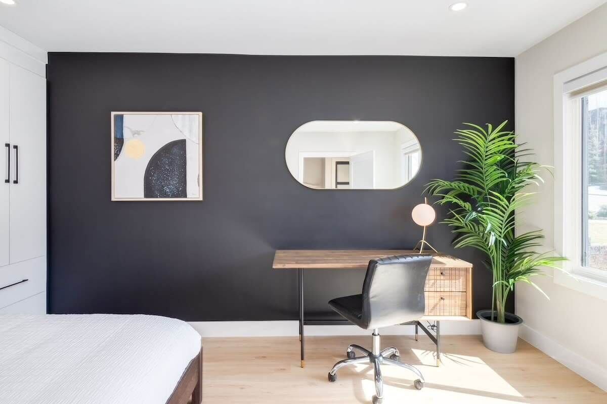 A modern bedroom with a bed, a desk, a chair, a potted plant, a mirror, and a painting on the wall.