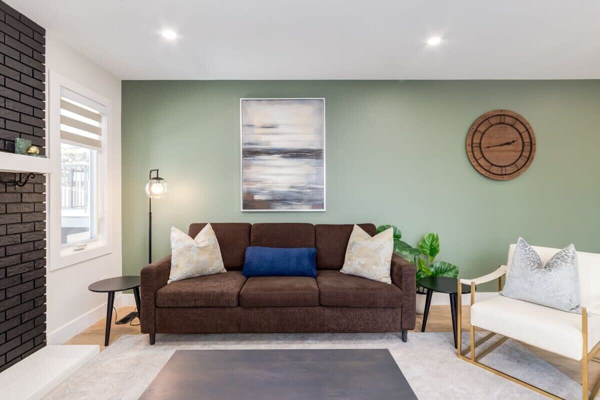 A living room with a brown sofa, a white armchair, a round wooden clock, a floor lamp, and a painting on the wall.
