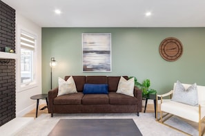 A living room with a brown sofa, a white armchair, a round wooden clock, a floor lamp, and a painting on the wall. - Pet Friendly 4BDR w/ Main Floor office/Bed (Calgary)