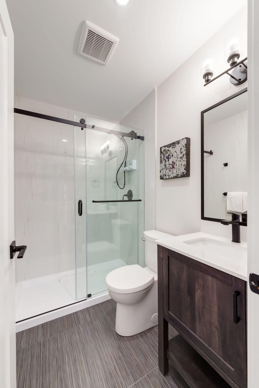 A modern bathroom with a glass shower enclosure, a white toilet, a dark wooden vanity, and a mirror with a light fixture.