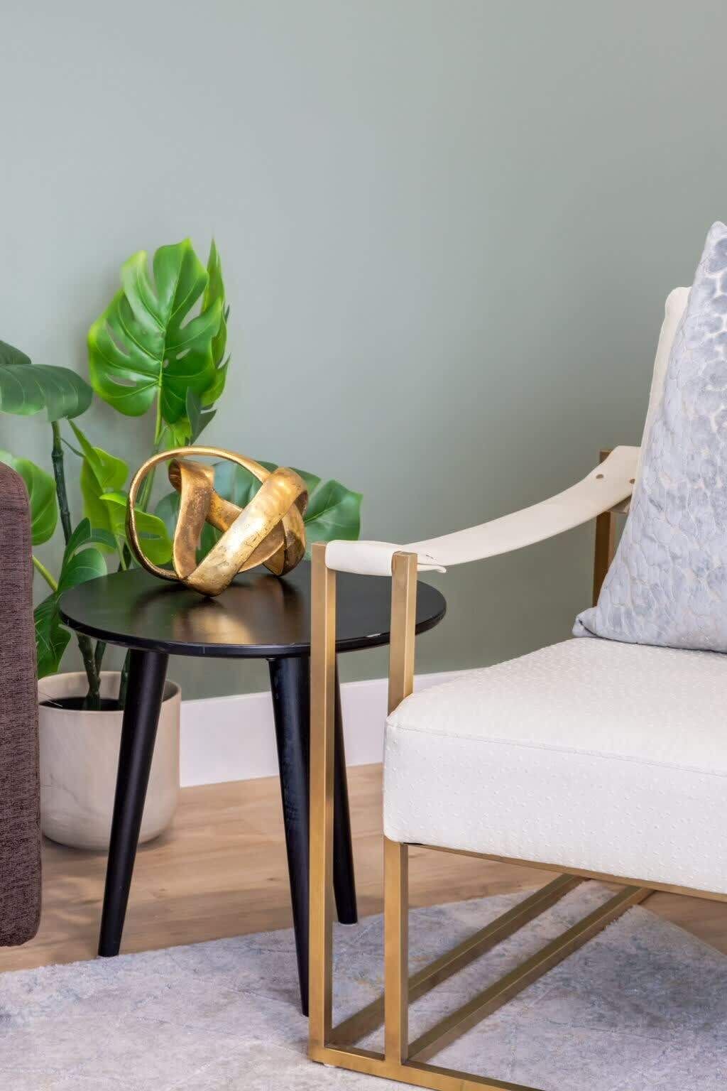 A room with a round black table, a white cushioned chair, a potted plant, and a grey wall.