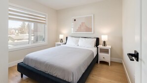 A bedroom with a bed, two bedside tables, a window with blinds, a wall-mounted artwork, and a door with a handle. - Pet Friendly 4BDR w/ Main Floor office/Bed (Calgary)