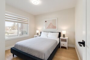 A bedroom with a bed, two bedside tables, a window with blinds, a wall-mounted artwork, and a door with a handle. - Pet Friendly 4BDR w/ Main Floor office/Bed (Calgary)