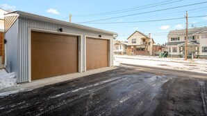 Un garaje residencial con dos puertas grandes, un camino de entrada y casas al fondo.