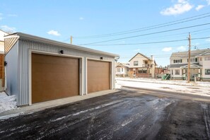 A residential garage with two large doors, a driveway, and houses in the background. - Free Parking, Pet Friendly, 3 BDR, Prime Location (Calgary)
