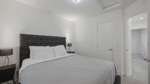 A modern bedroom with a large bed, a nightstand, a lamp, and a door leading to another room.