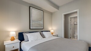 Une chambre à coucher bien aménagée, avec un grand lit, deux tables de chevet avec des lampes, une œuvre d’art abstraite encadrée au-dessus du lit, et une porte donnant accès à une salle de bain.