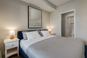 Une chambre à coucher bien aménagée, avec un grand lit, deux tables de chevet avec des lampes, une œuvre d’art abstraite encadrée au-dessus du lit, et une porte donnant accès à une salle de bain.
