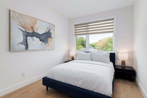 A bedroom with a bed, two bedside tables with lamps, a window with blinds, and a large abstract painting on the wall.