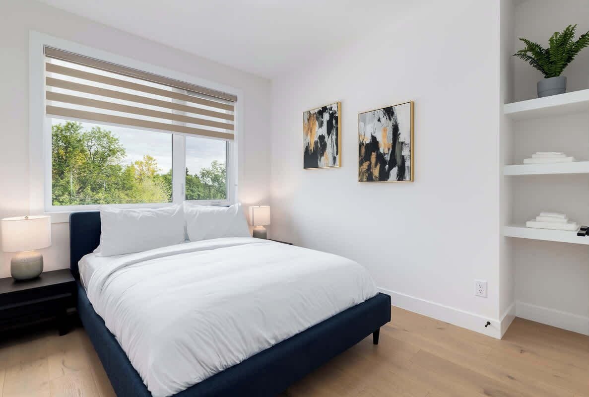 Ein modernes Schlafzimmer mit einem großen Bett, einem Fenster mit Jalousien, zwei abstrakten Gemälden und einer weißen Regaleinrichtung.