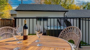 Uma mesa de madeira com quatro copos de vinho e uma garrafa, com um cercado branco e uma casa ao fundo.