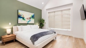 Quarto com cama, mesa de cabeceira, TV na parede e janela com persianas.