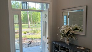 Un foyer avec une porte blanche donnant sur un patio couvert, un tapis gris, une console en bois ornée d’un vase de fleurs et un miroir fixé au mur.