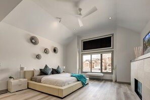 Une chambre à coucher avec un grand lit, une table de chevet, une commode, un ventilateur de plafond, une grande fenêtre et une cheminée.