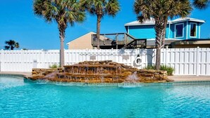 Piscina con cascada de roca, rodeada de palmeras y una cerca blanca.