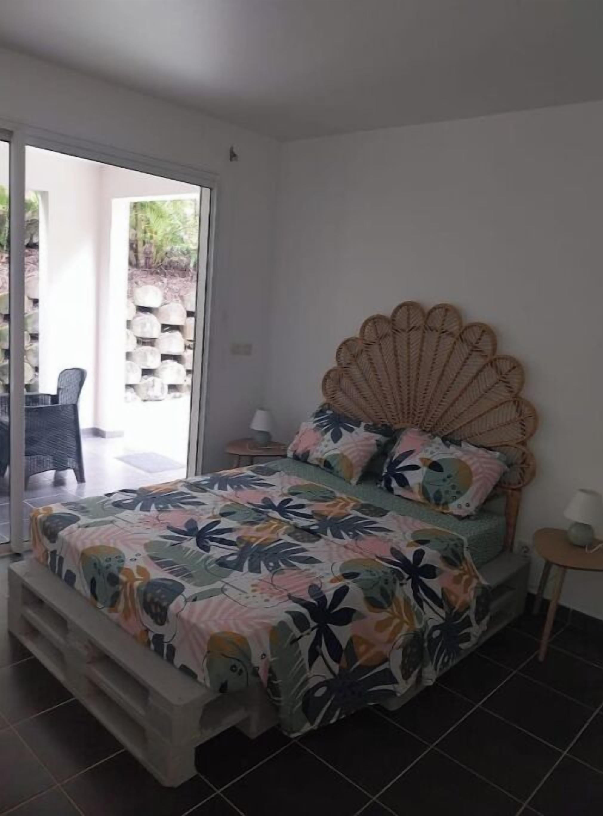 Een slaapkamer met een bed, een rieten hoofdbord, een raam en uitzicht op een tuin.