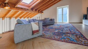 Una stanza in stile loft con soffitto alto, travi in legno, un'ampia vetrata a soffitto, un divano grigio, un tappeto decorato e una porta scorrevole in vetro.