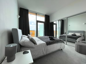 Quarto de hotel moderno com uma cama grande, mesa de cabeceira, espelho e vista para a cidade pela janela.