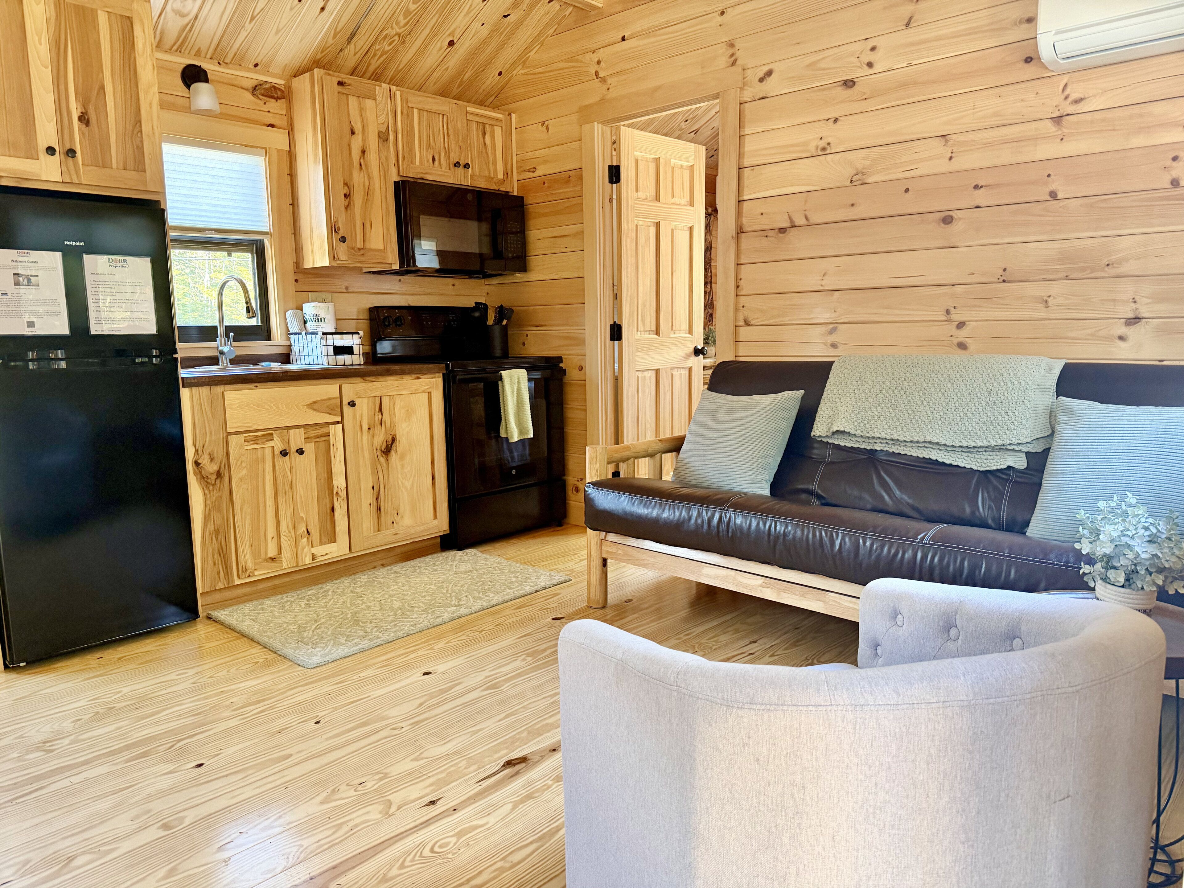 A wooden cabin interior with a kitchen, a black refrigerator, a microwave, a stove, a couch, and a chair.