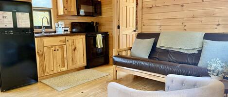 A wooden cabin interior with a kitchen, a black refrigerator, a microwave, a stove, a couch, and a chair.