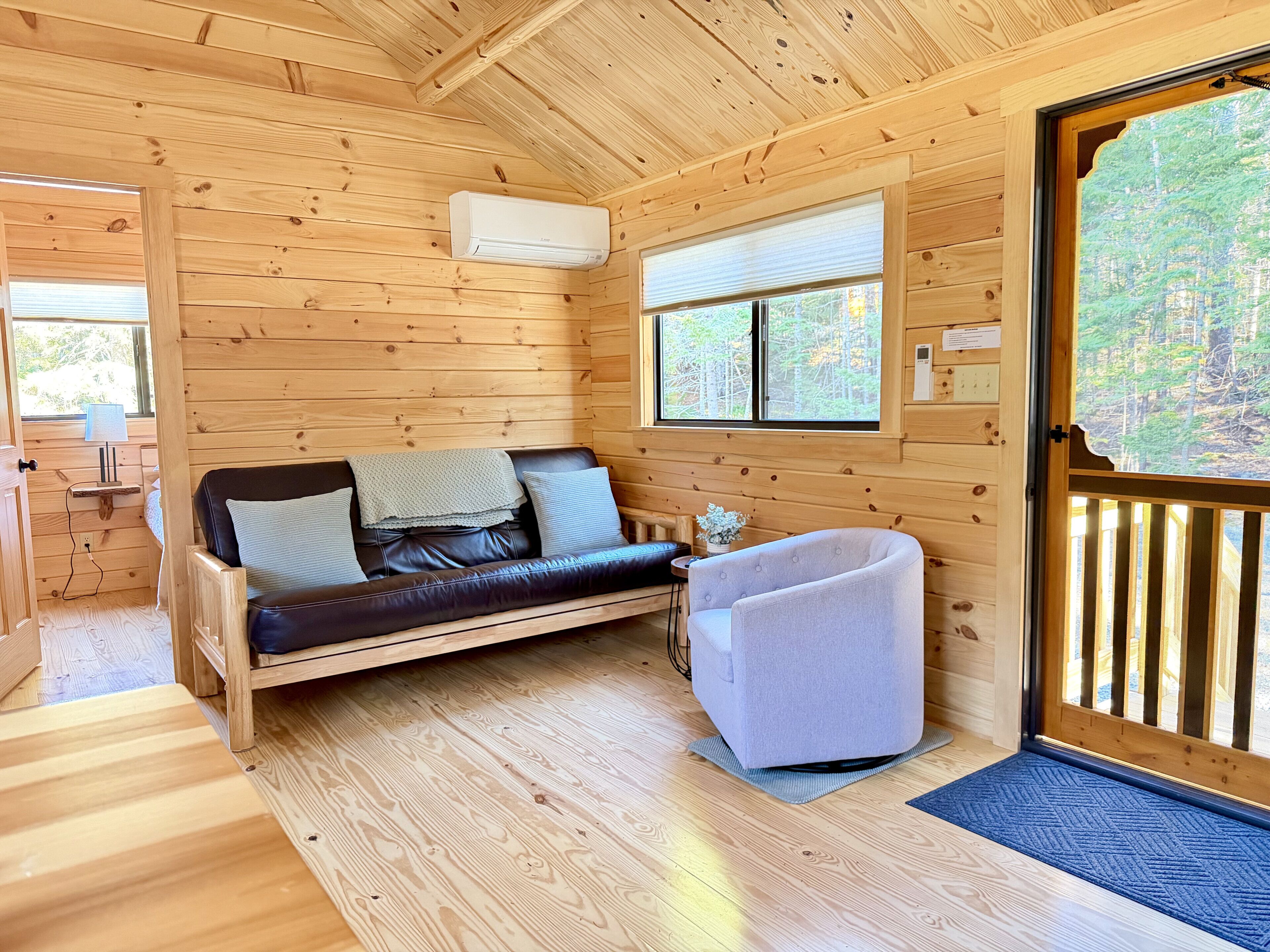 L’intérieur d’un chalet en bois, avec un canapé en cuir, un petit fauteuil, une fenêtre avec des stores et une porte donnant sur l’extérieur.