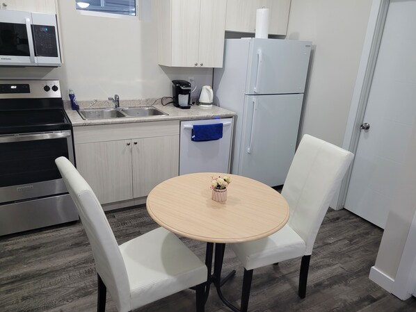 Cocina compacta con gabinetes blancos, estufa de acero inoxidable, refrigerador y un lavamanos. Hay una pequeña mesa con un florero y dos sillas blancas.