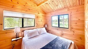 Une chambre dans une cabane en bois, avec un lit, deux fenêtres et une table de chevet avec une lampe.