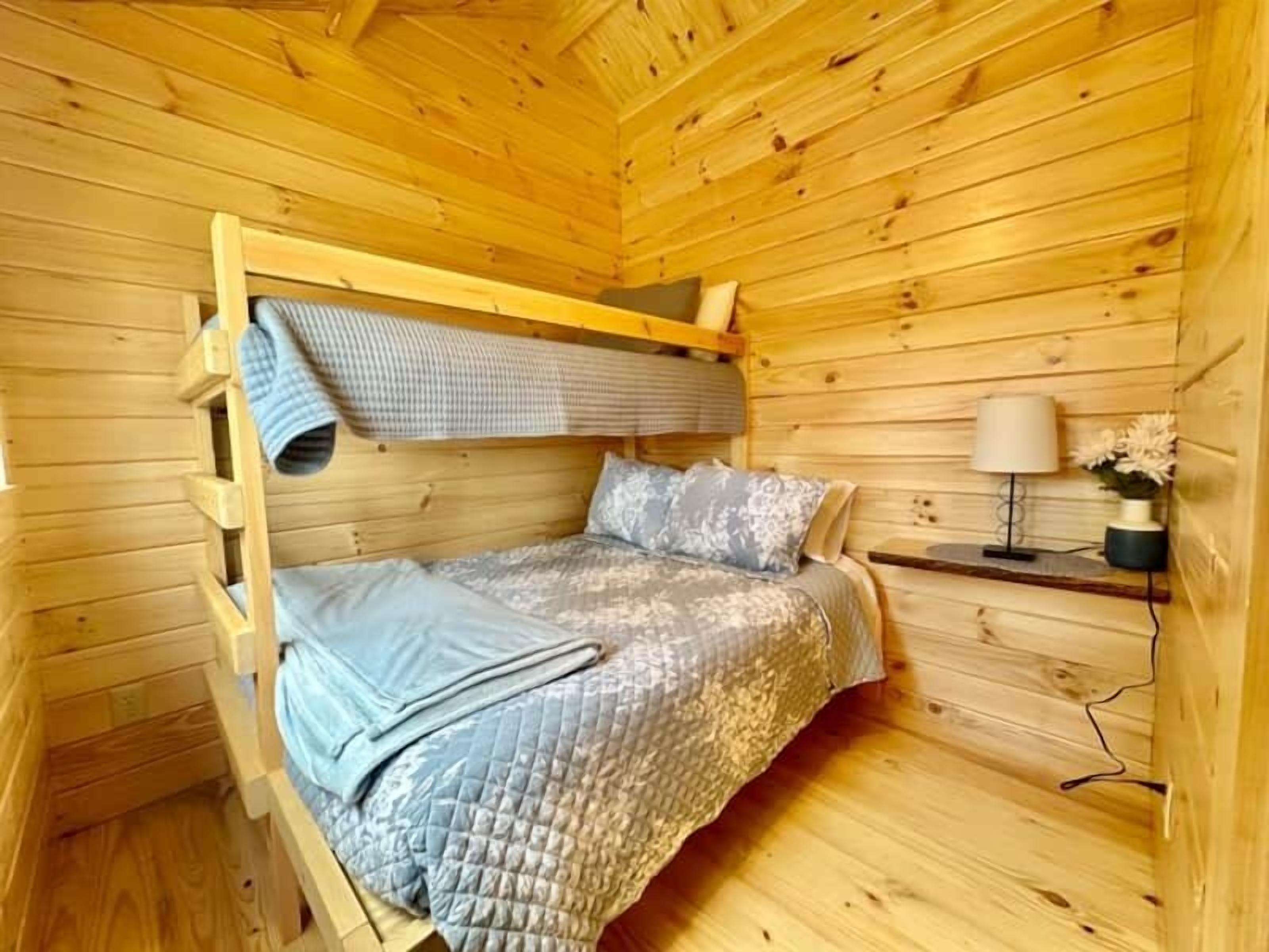 A wooden cabin room with a bunk bed, a bedside table with a lamp, and a small shelf with flowers.