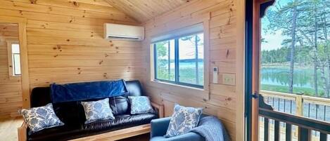 A cozy cabin interior with a wooden ceiling, a sofa, a chair, and a large window offering a view of the outdoors.