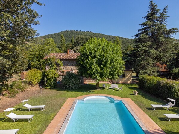 A swimming pool with lounge chairs, a stone building, and a forested hill in the background.
