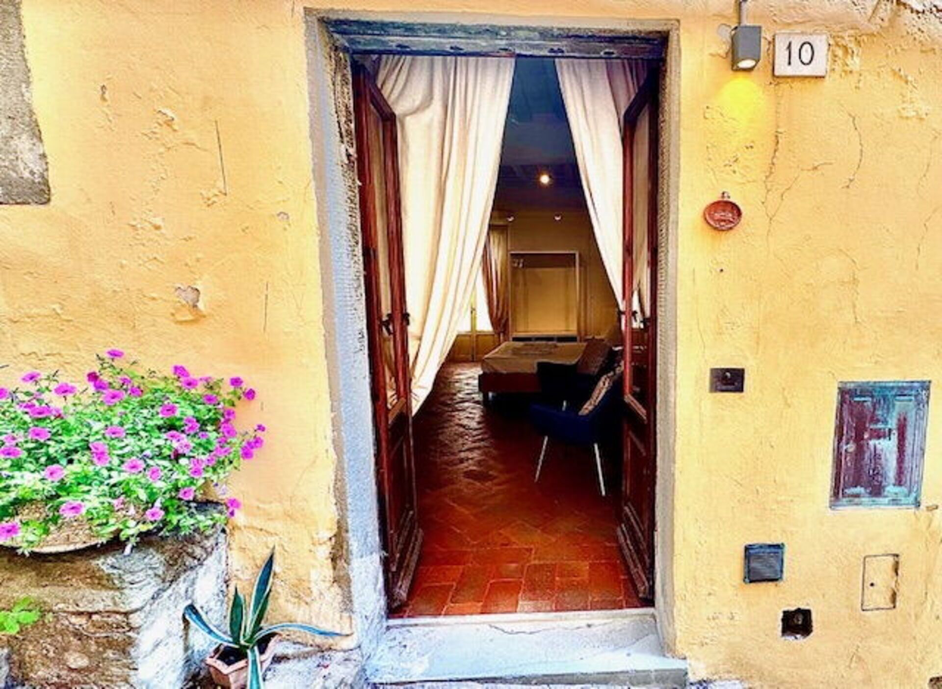 A narrow entrance with a bed and a chair visible inside, a number '10' sign, and a flower box with pink flowers.