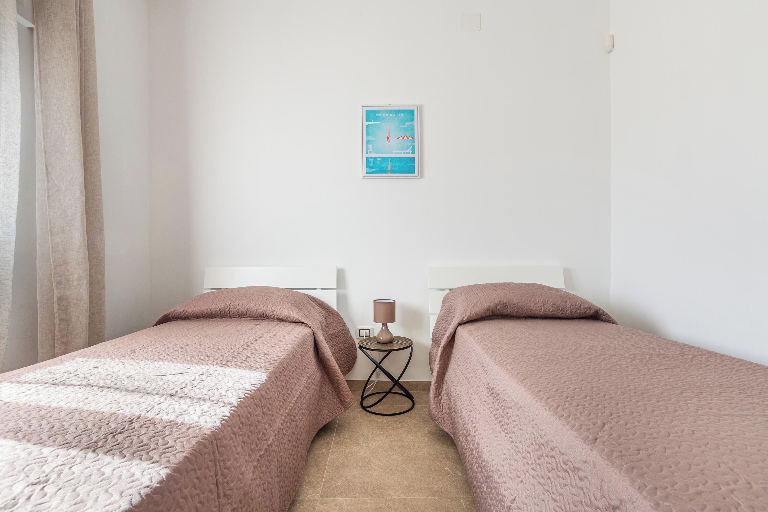 A small, clean room with two beds, a small table with a lamp, and a framed picture on the wall.