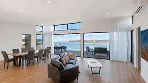 A spacious living area with a wooden floor, a sofa, a coffee table, and a dining table with chairs. Large windows offer a view of the sea and a crane. - SouthWind (Merimbula)