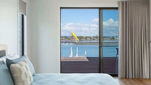 A bedroom with a large bed, a view of a body of water, and a balcony with a chair. - SouthWind (Merimbula)