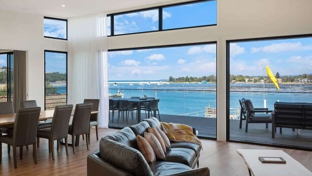 A spacious living area with a large window overlooking a marina, a dining area with chairs, and a sofa with cushions. - SouthWind (Merimbula)